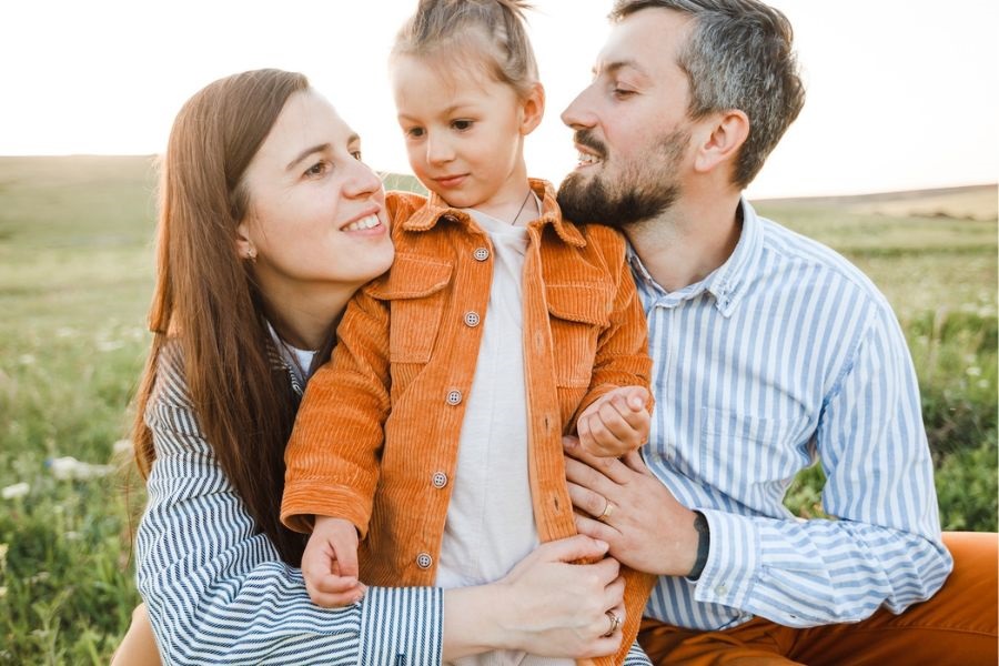 A man and woman sit on a grassy field, smiling at their daughter who plays nearby, enjoying a sunny day together